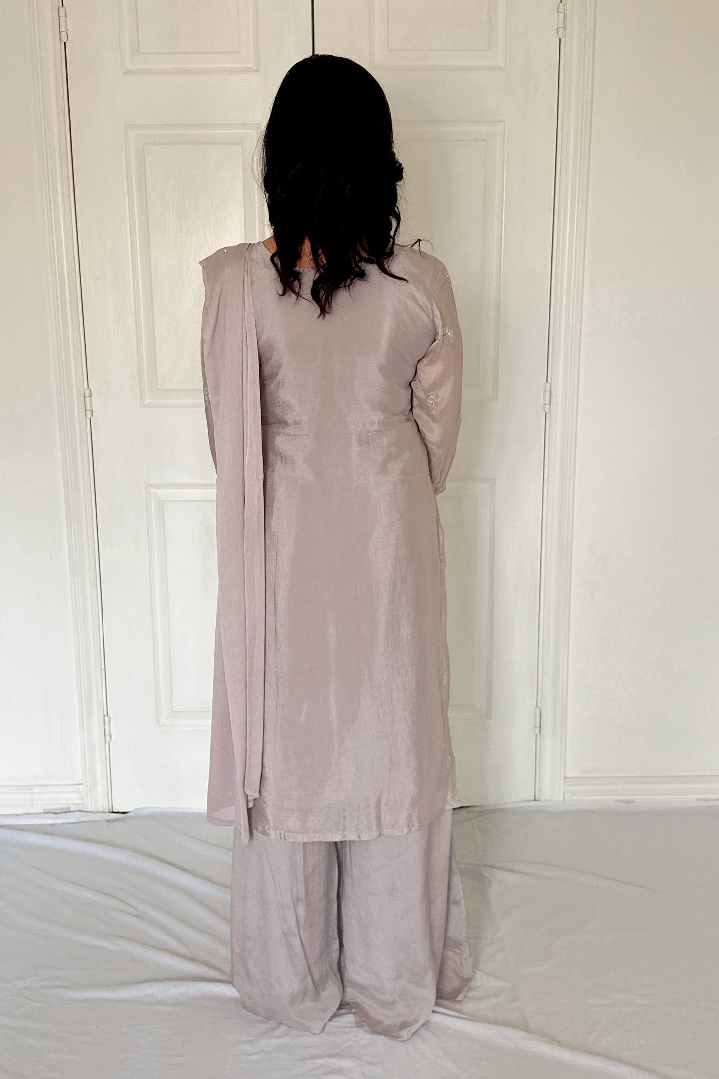 Person wearing a light gray outfit standing against a white paneled wall.